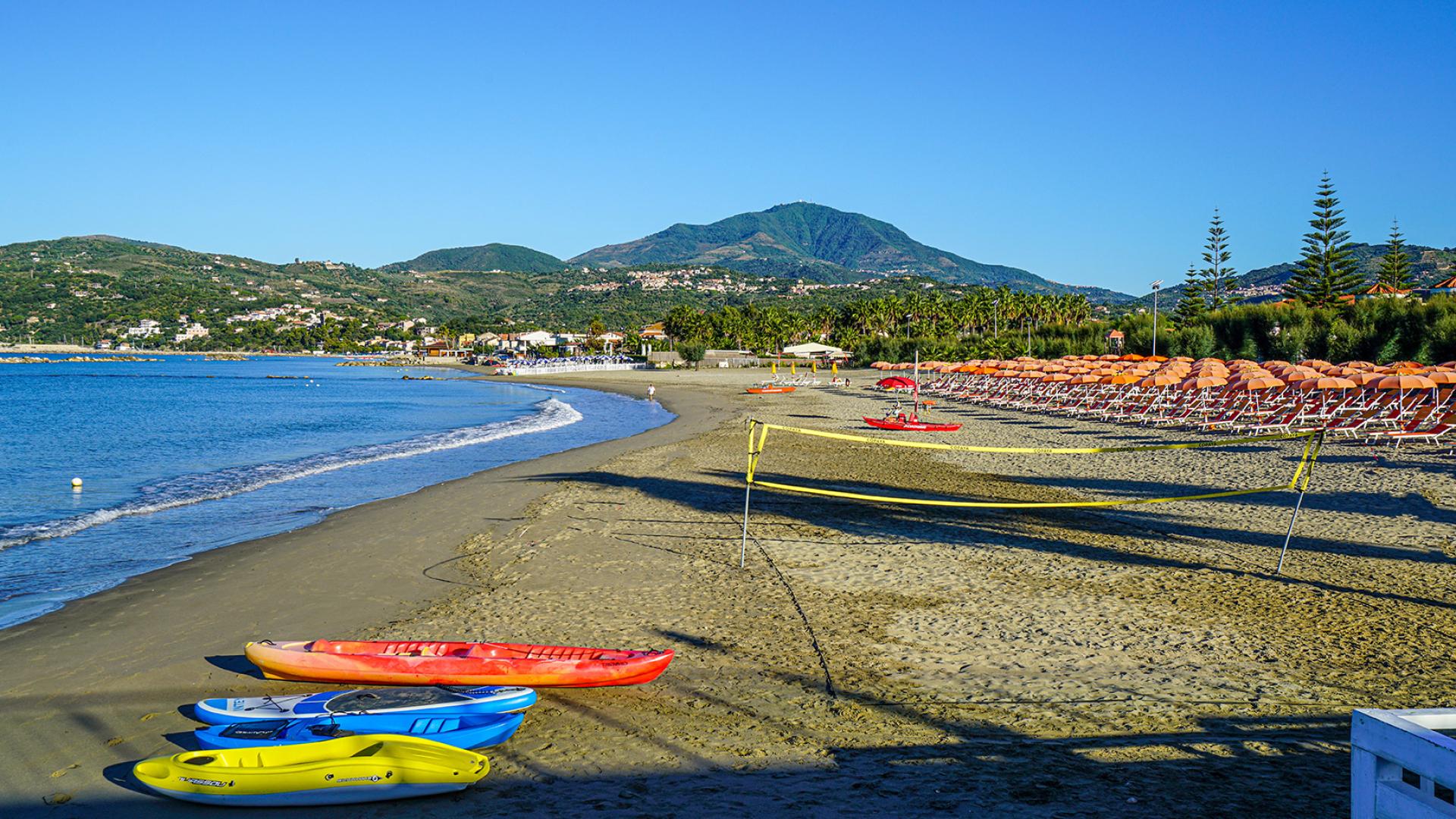 Spiaggia con canoe colorate e ombrelloni arancioni, circondata da montagne e mare calmo.