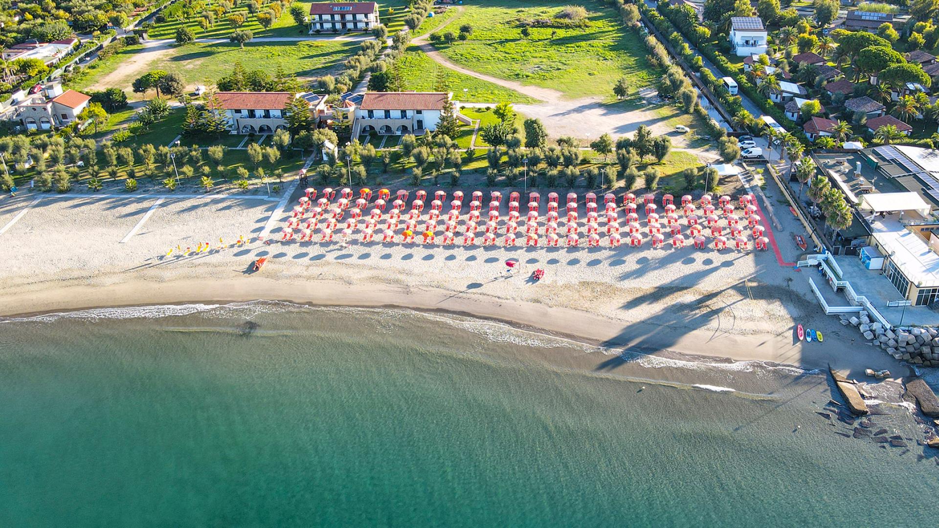 Spiaggia con ombrelloni rossi, mare calmo e strutture turistiche immerse nel verde.