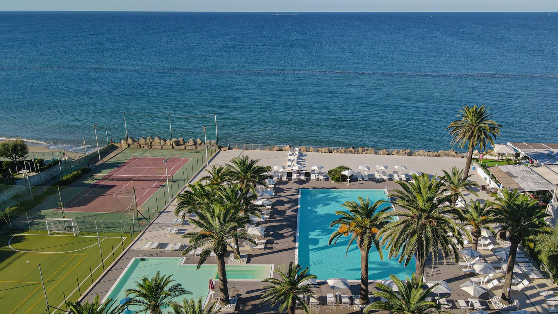 Vista su piscina, campi sportivi e mare con palme e lettini.