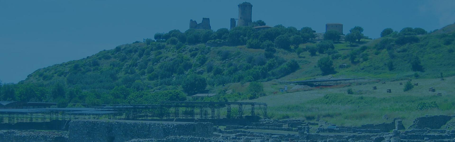 Rovine antiche su una collina verde con strutture storiche in lontananza.