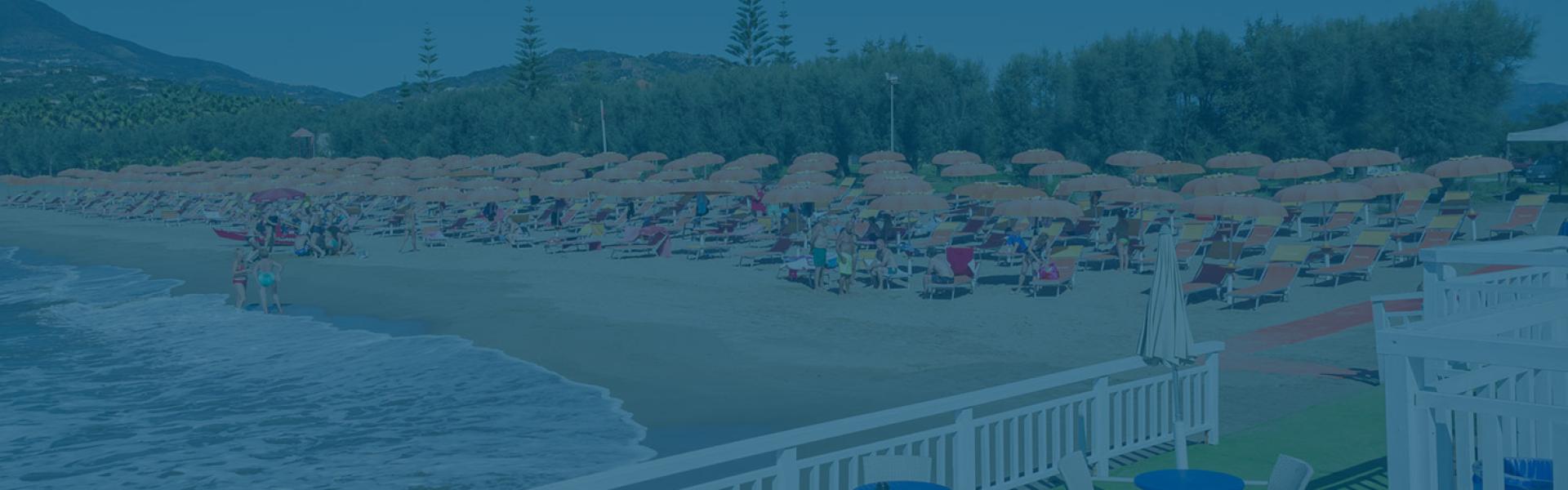 Spiaggia affollata con ombrelloni arancioni, mare calmo e colline sullo sfondo.