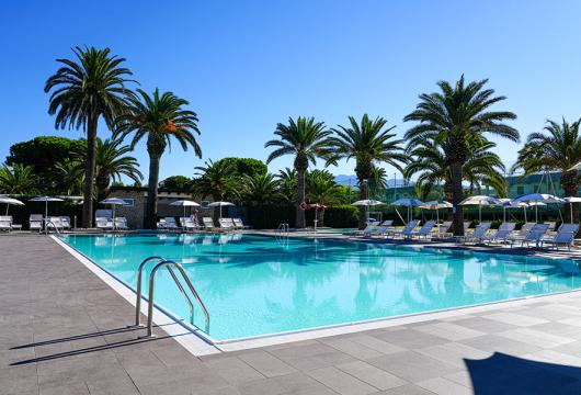 Piscina all'aperto con palme e lettini sotto un cielo sereno e soleggiato.