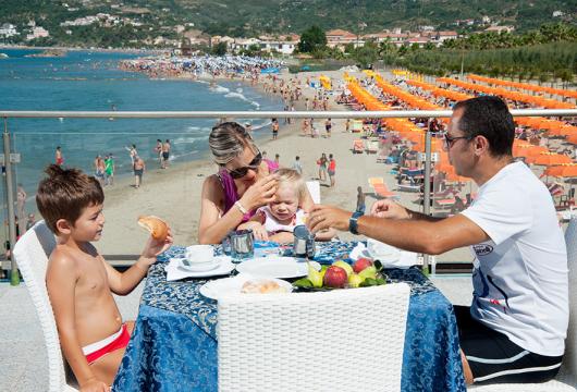 Famiglia fa colazione vista mare, spiaggia affollata e ombrelloni arancioni sullo sfondo.