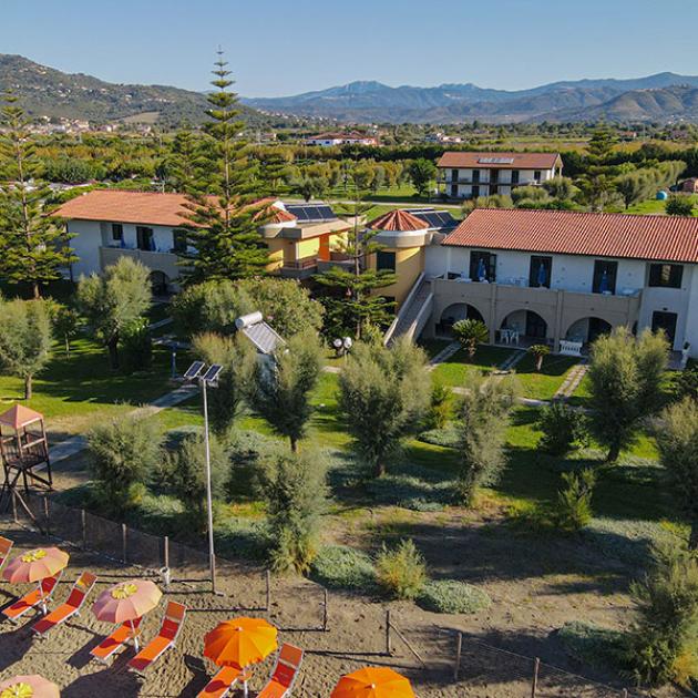 Resort con giardino, ombrelloni colorati, vista monti e tetti rossi.