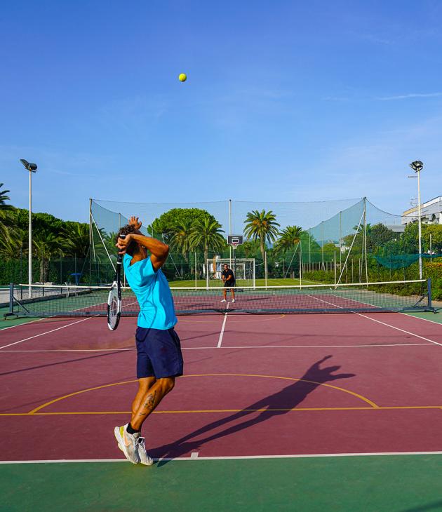 Giocatori di tennis su un campo all'aperto, circondato da palme e edifici.