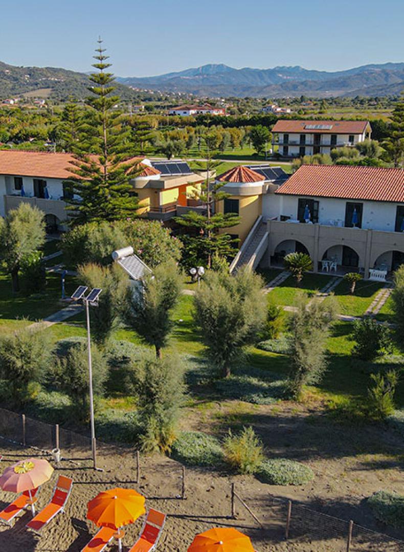 Vista aerea di un villaggio turistico con ombrelloni e giardini, circondato da montagne.