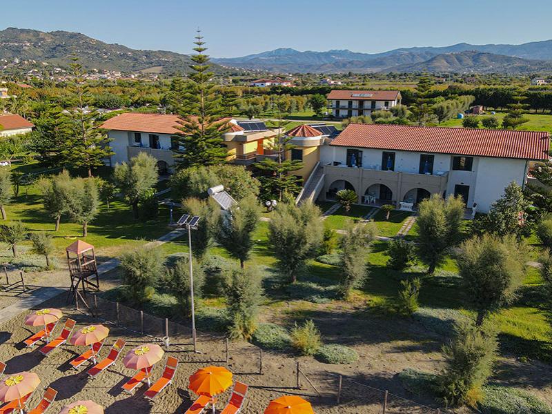 Resort con giardino, ombrelloni colorati, vista monti e tetti rossi.