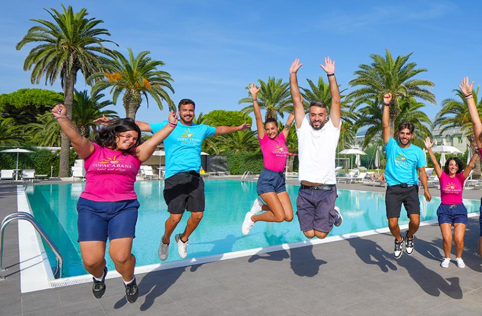 Gruppo di persone salta felice davanti a una piscina, palme sullo sfondo.