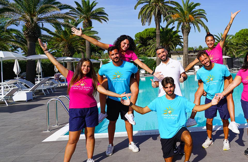 Gruppo di persone sorridenti in abbigliamento estivo vicino a una piscina con palme.