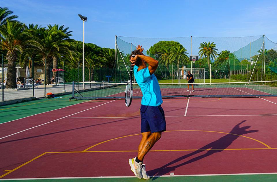 Due persone giocano a tennis su un campo all'aperto con palme sullo sfondo.