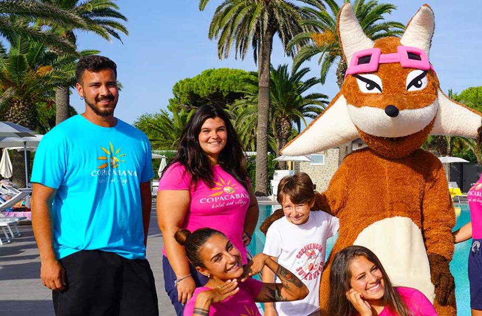 Gruppo di persone sorridenti con una mascotte vicino a una piscina, palme sullo sfondo.