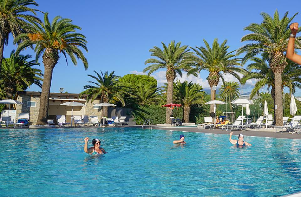 Piscina con palme, persone che nuotano e ragazza sorridente in maglietta rosa.