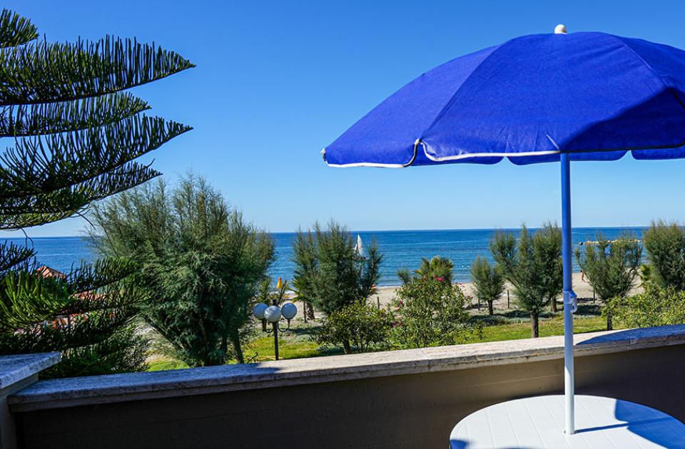 Vista mare con ombrellone blu, alberi e cielo sereno.