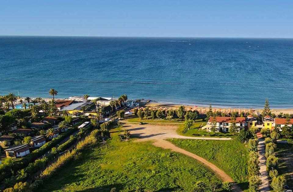 Vista aerea di una spiaggia con ombrelloni e edifici circondati da vegetazione.