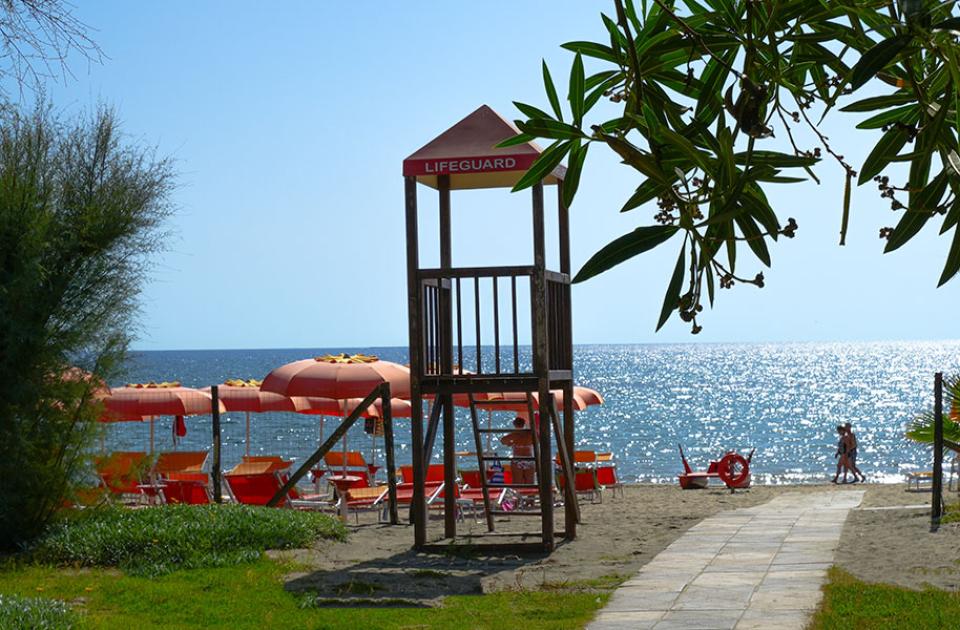 Spiaggia con ombrelloni arancioni, torretta bagnino e doccia, vista mare scintillante.