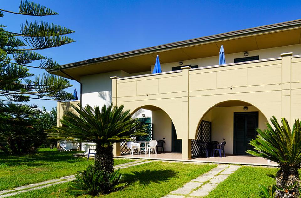 Villetta con archi, giardino curato e ombrelloni blu, in ambiente soleggiato.