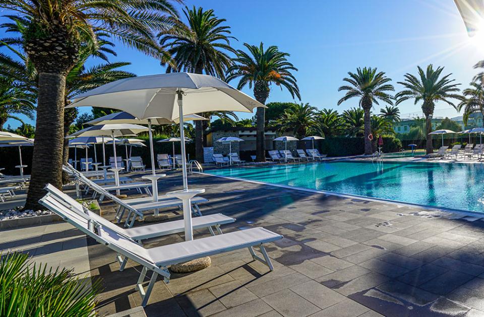 Piscina all'aperto con lettini, ombrelloni e palme sotto il sole.