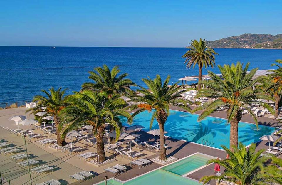 Piscina con palme e vista mare, lettini e ombrelloni su una terrazza soleggiata.