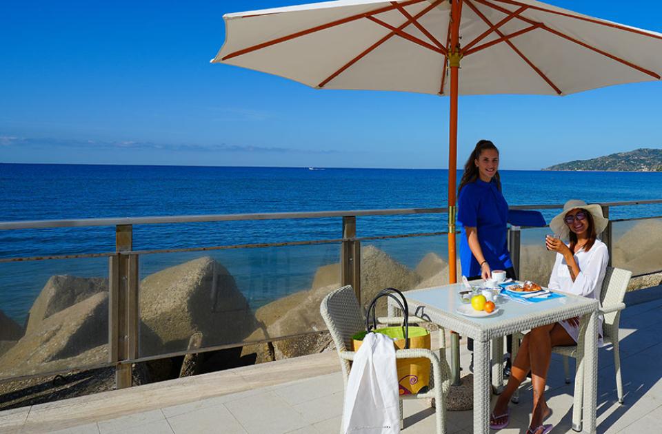 Colazione all'aperto con vista mare, sotto un ombrellone su una terrazza soleggiata.