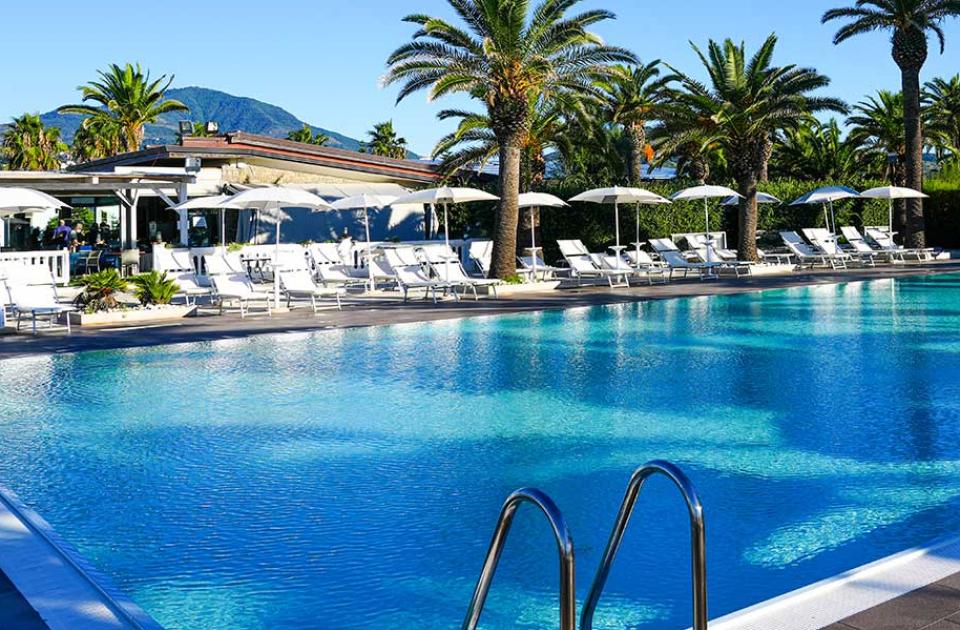 Piscina all'aperto con lettini, ombrelloni e palme, immersa in un ambiente tropicale.