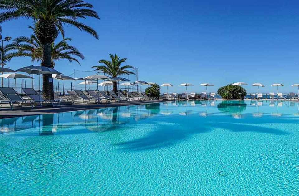 Piscina all'aperto con palme, lettini e ombrelloni sotto un cielo azzurro.