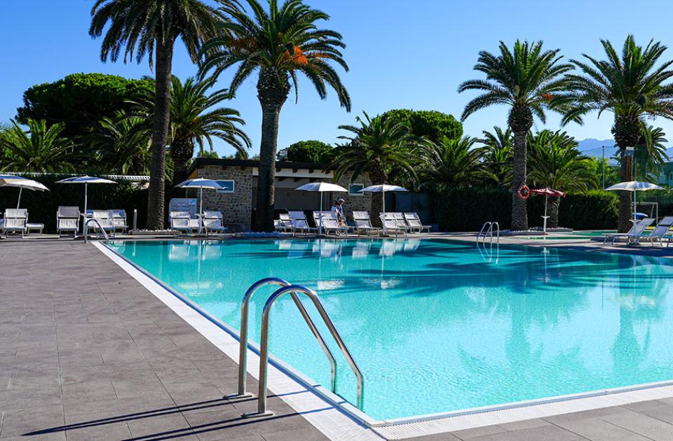 Piscina all'aperto con lettini, ombrelloni e palme in un ambiente soleggiato.