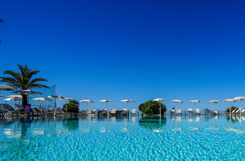 Piscina con ombrelloni e palme, sotto un cielo blu intenso.