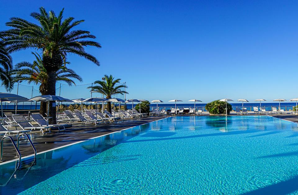 Piscina all'aperto con lettini, ombrelloni e vista mare, circondata da palme.