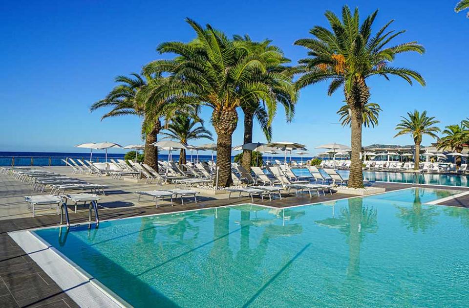 Piscina con palme e lettini, vista mare e cielo azzurro.