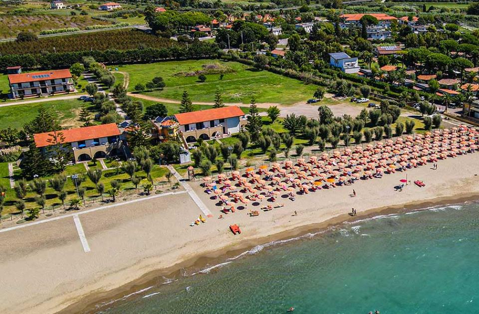 Spiaggia con ombrelloni, mare cristallino e villette immerse nel verde, vista aerea.