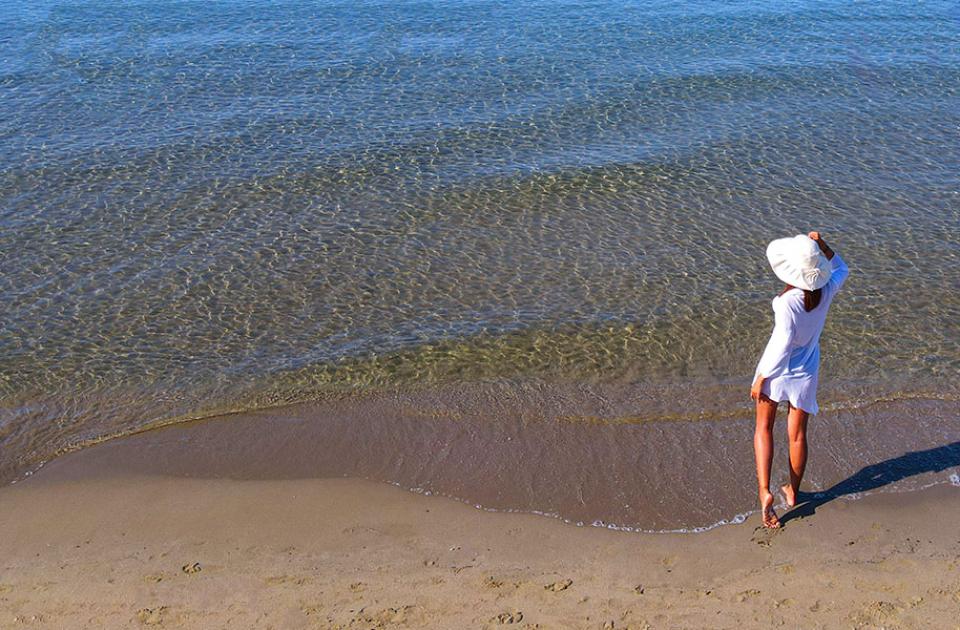 Donna in spiaggia con cappello bianco, guarda il mare.