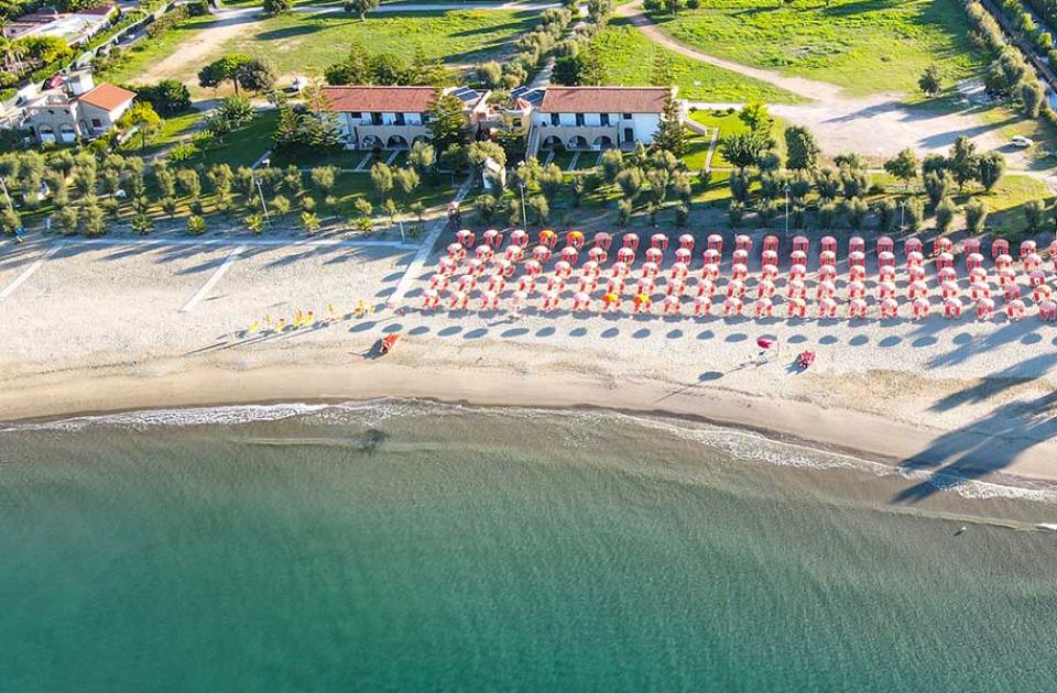 Spiaggia con ombrelloni rossi e bianchi, vista dall'alto, vicino a edifici e vegetazione.