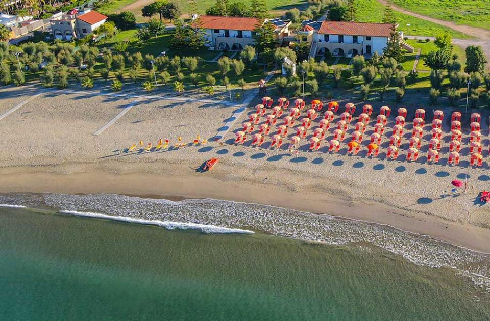 Spiaggia con ombrelloni rossi e arancioni, vista dall'alto, vicino a edifici e vegetazione.