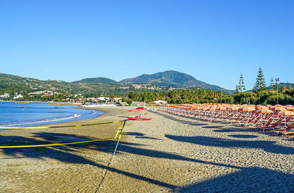 Spiaggia sabbiosa con ombrelloni arancioni, mare calmo e montagne sullo sfondo.