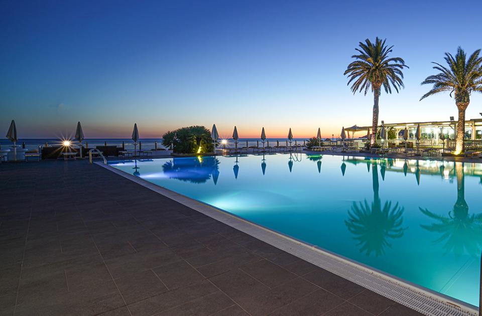 Piscina illuminata al tramonto con palme e vista sul mare.